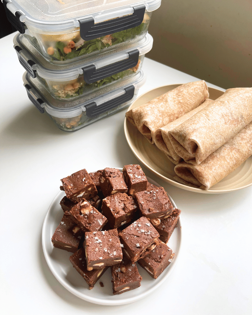 glass meal prep containers with salad, 4 wraps on a plate, and protein fudge on a white plate, all on a white surface