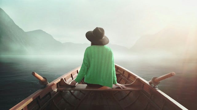 A woman in a boat on calm water, with her back to the camera, surrounded by mist and distant mountains, evoking peace and quiet anticipation.