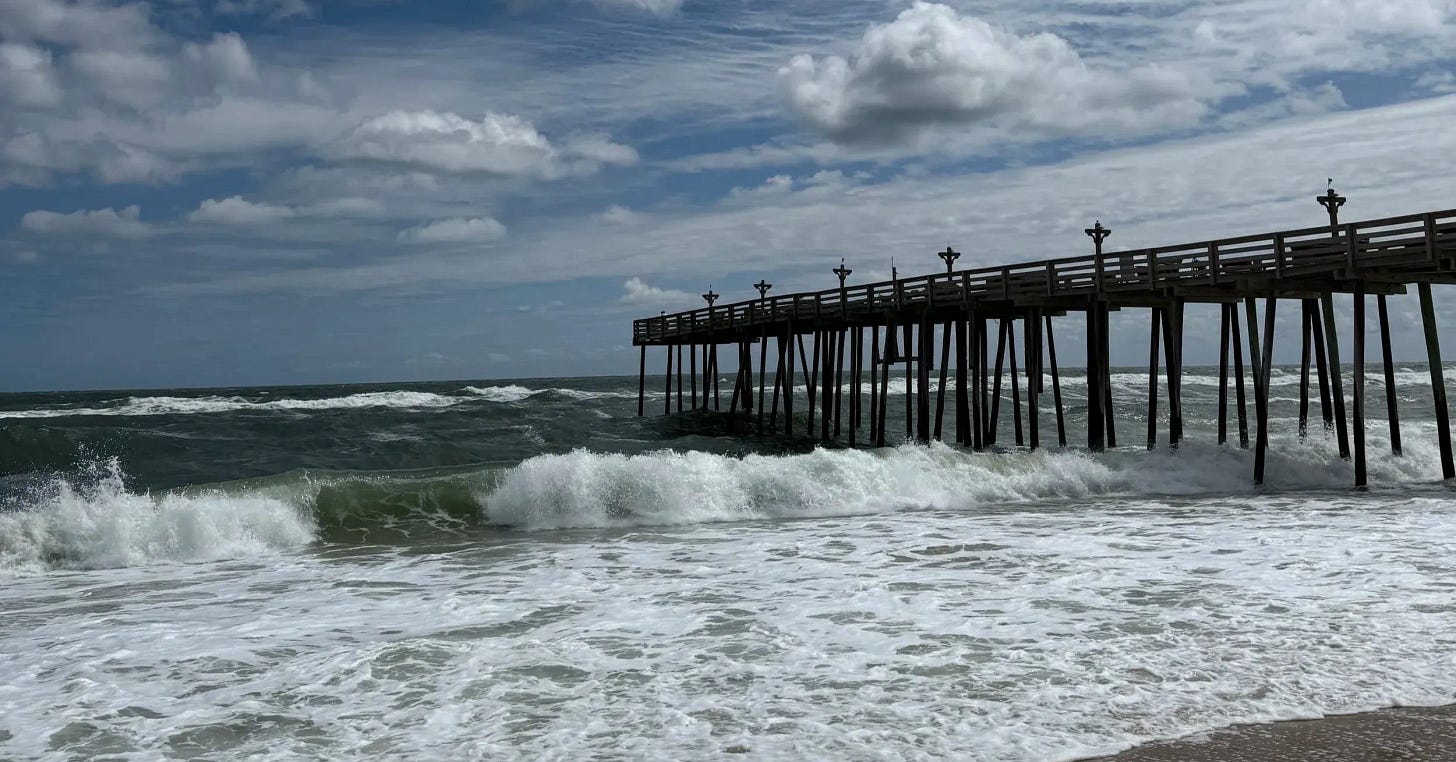 Ocean waves breaking beside a wooden pier under a bright coastal sky with scattered clouds