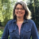 A photo of a white woman with shoulder-length grey hair. She is wearing a red and white striped t-shirt and a denim jacket