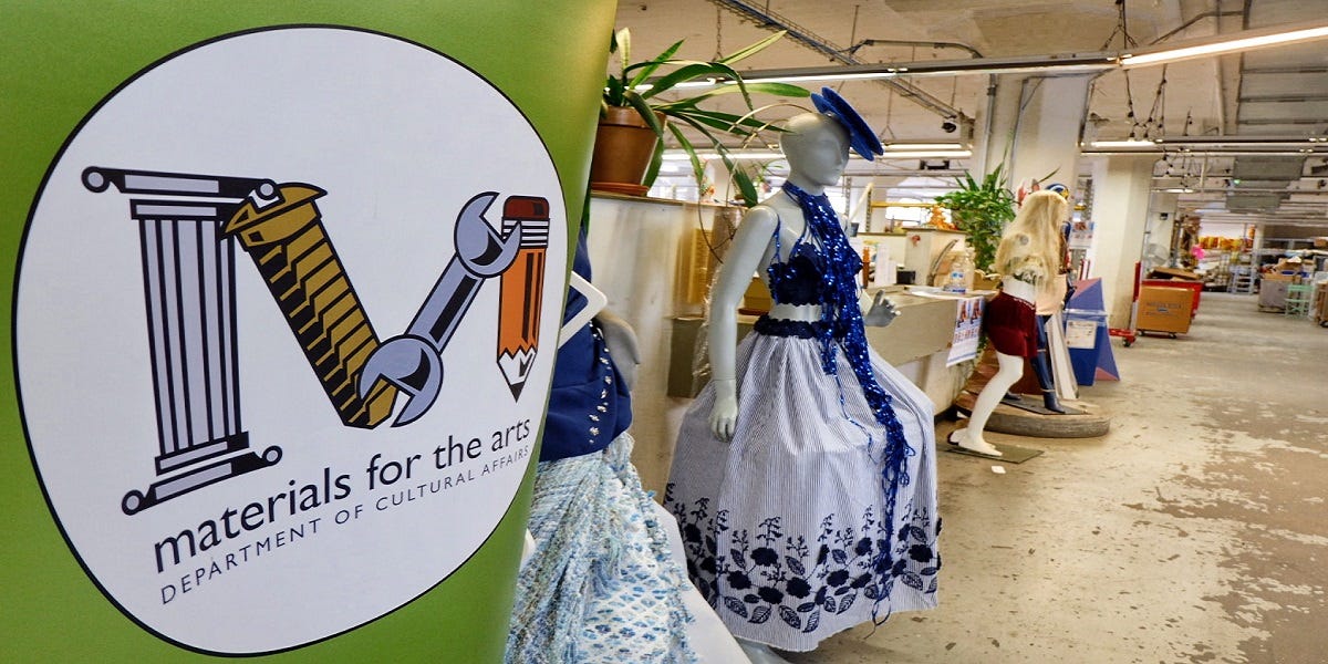The logo for materials for the arts which is a letter M made from a column, a screw, a wrench and a pencil. In the background is the warehouse with mannequins dressed in homemade costumes, plants, and rows of supplies