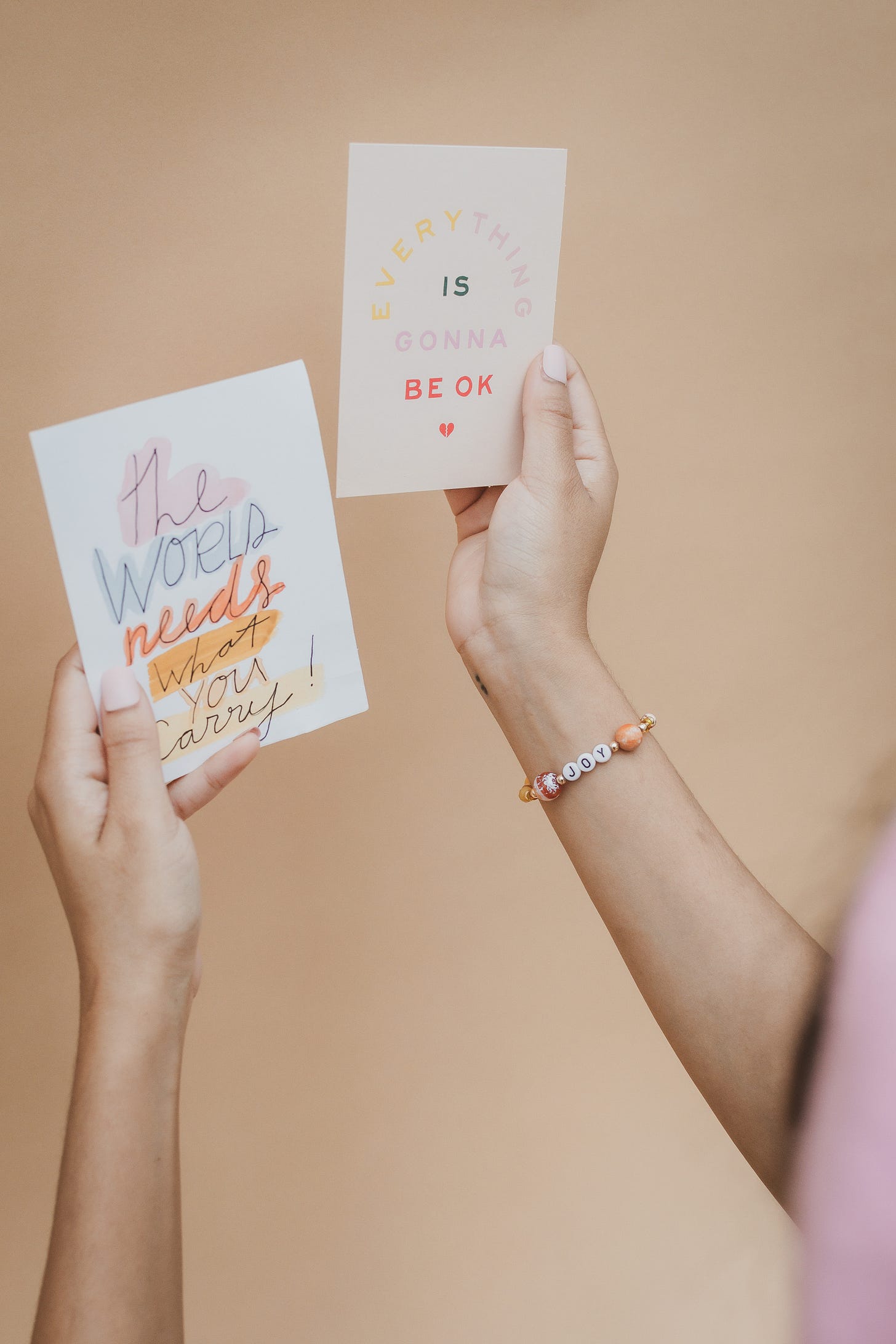 Kiana is holding up two greeting cards. One says the world needs what you carry and the other says everything is going to be okay. She is wearing a beaded bracelet that has the word joy on it