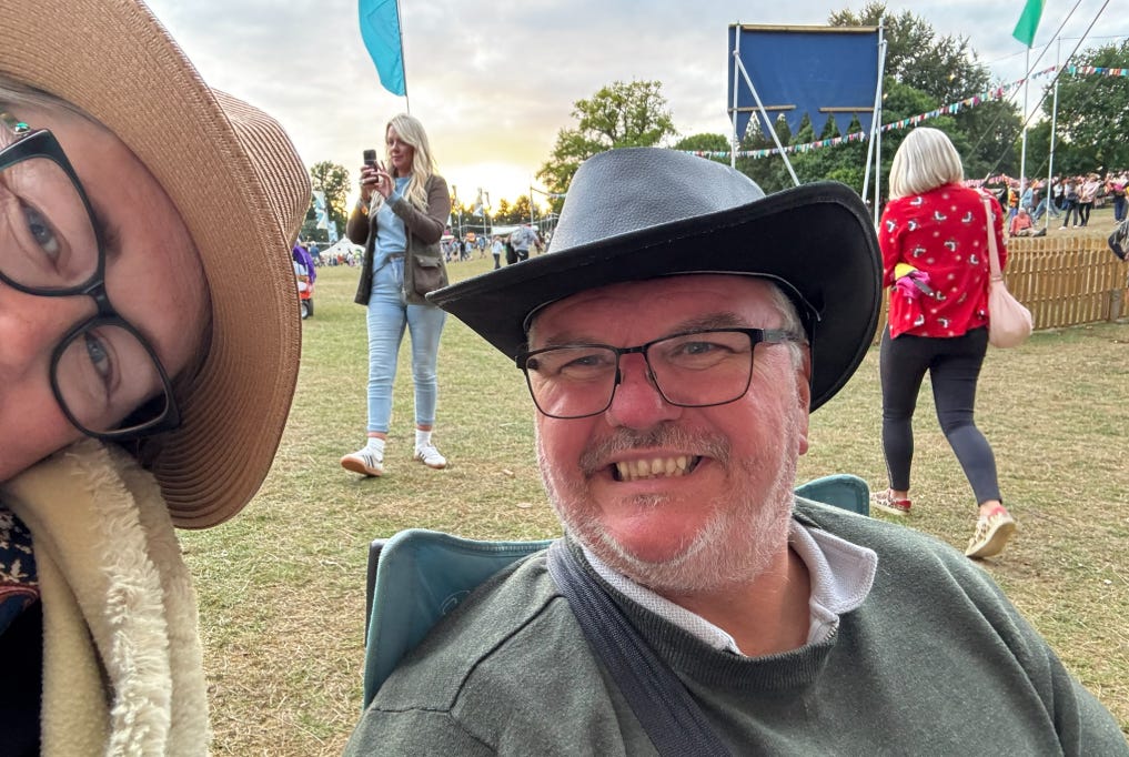 Two people sitting together at an outdoor festival. Both are wearing hats and glasses. Peter is smiling in a black cowboy‑style hat, and I am beside him in a tan hat and shawl. Colourful flags hang overhead and people are gathered in the background on a sunny day.