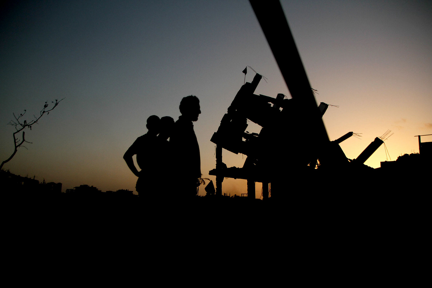 Forte contraluz que siluetea a 3 homes fronte a edificios convertidos en ruínas en Gaza. Anoitece polo que as cores son na súa maioría escuros e dourados.