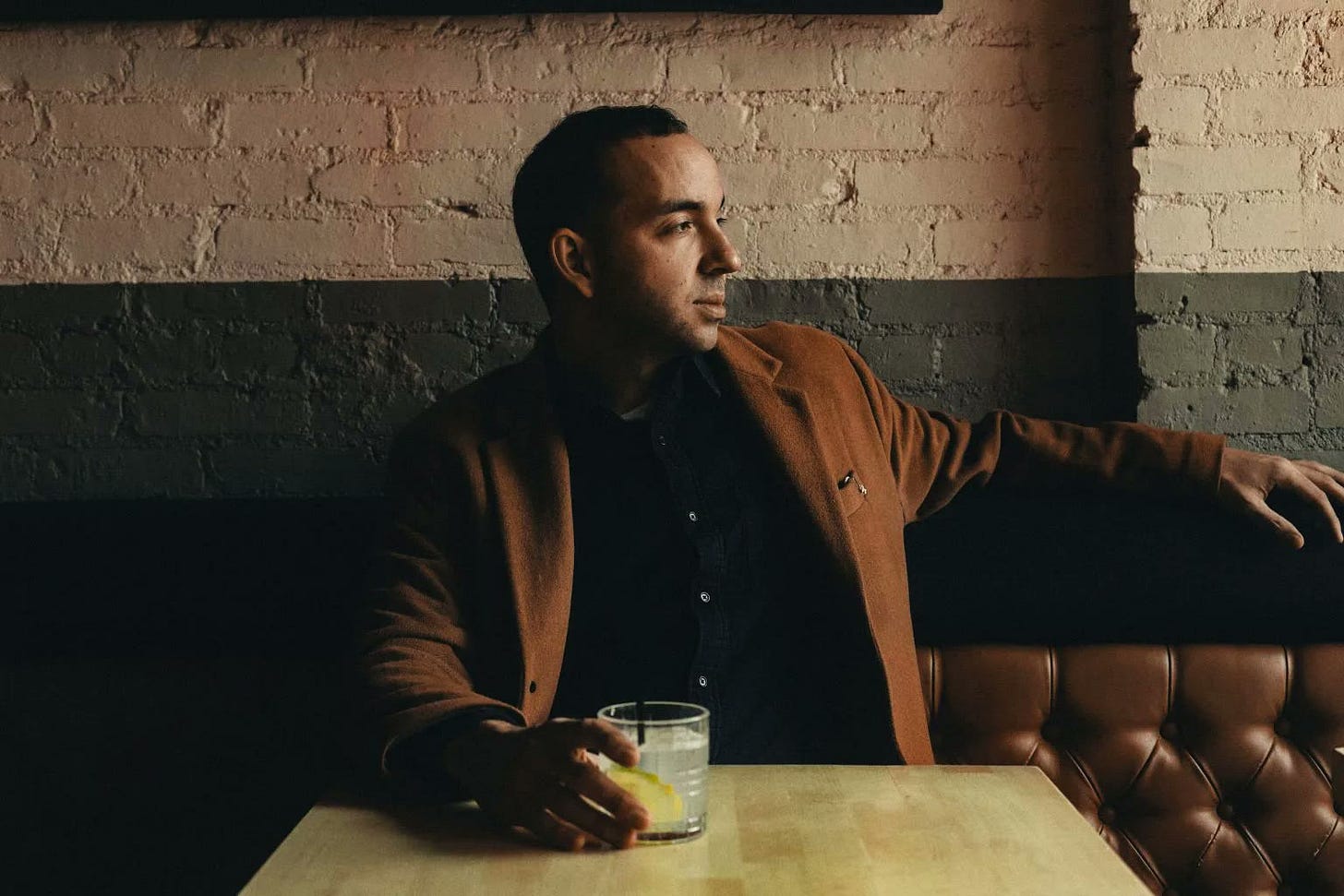 King Klave, Amaury Acosta, in a camel coat at a booth table against a brick wall in Hell's Kitchen.