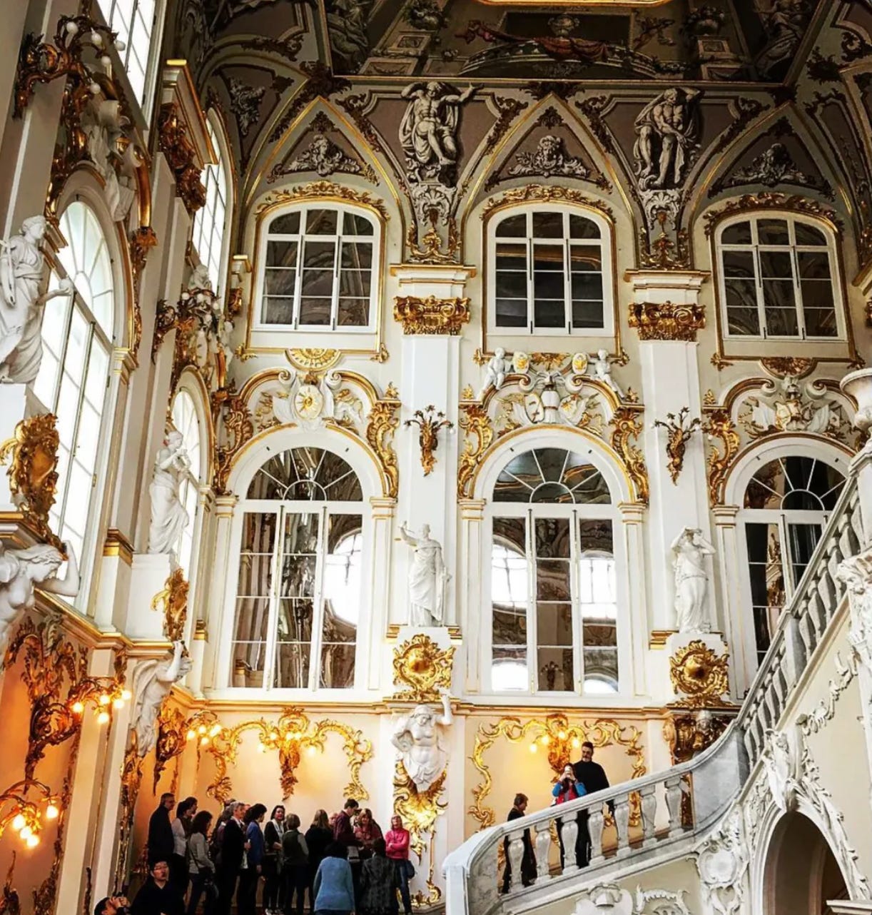 The ornate Jordan Staircase inside the Hermitage Museum, featuring sweeping white marble stairs, elaborate gold rococo decoration, classical statues in niches, and crowds of tourists ascending beneath painted ceiling frescoes.