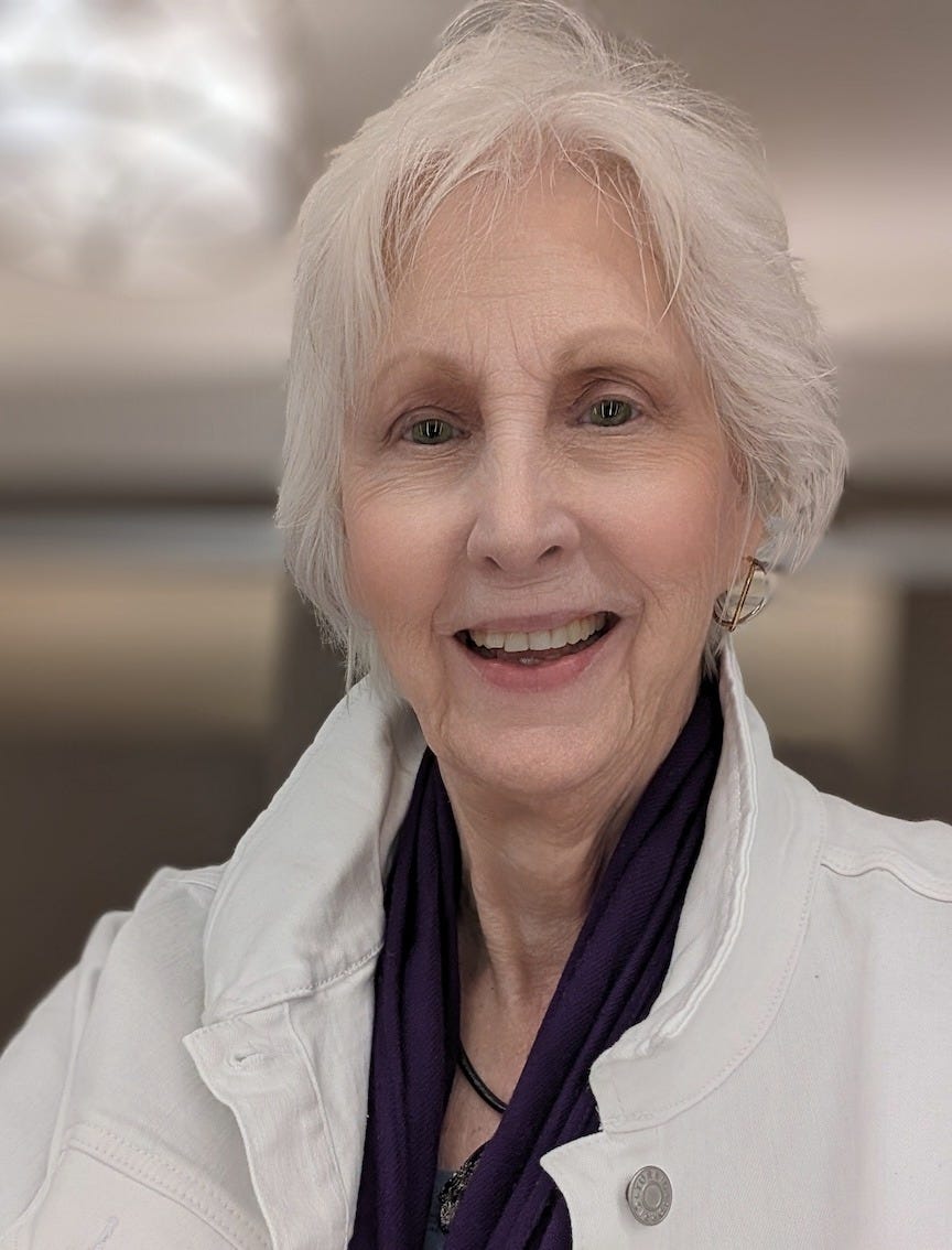 Selfie of Donna Blevins, the author, at 75, wearing a white jean jacket with a purple scarf and a gold earring showing on her left ear. She is proud of her well-earned wrinkles and her short, white hair. The background is blurred.
