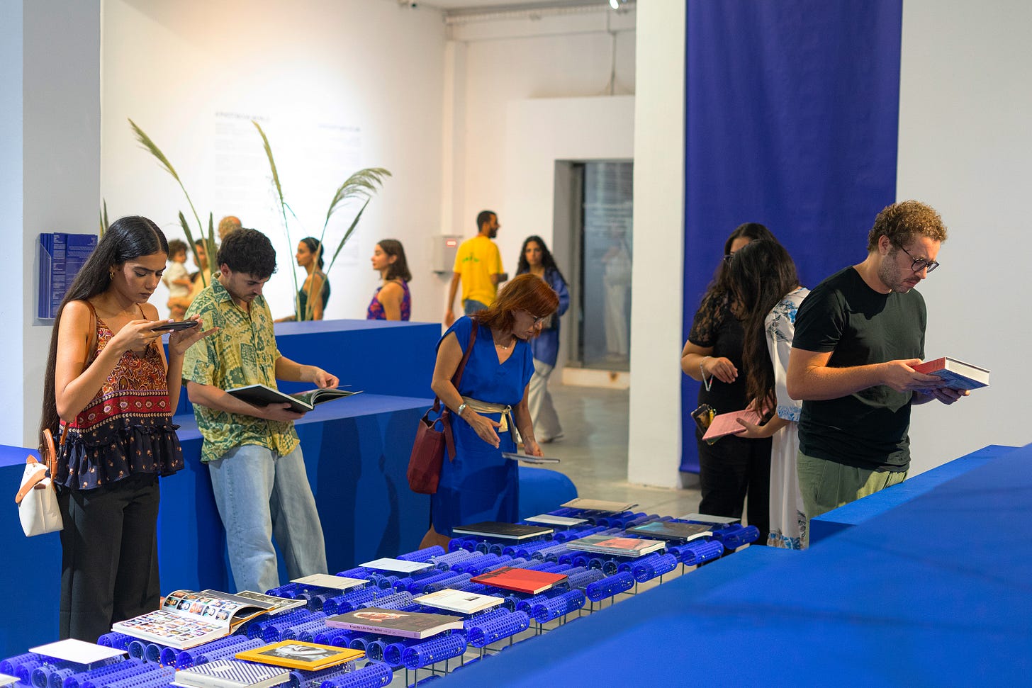 An image of a gallery with a blue table and benches. On the table, a collection of photography books are being browsed by people. Some are reading the books, one is staking a photograph of a spread. In the background, more people are checking the rest of the exhibition. An image of a gallery with a blue table and benches. On the table, a collection of photography books are being browsed by people. Some are reading the books, one is staking a photograph of a spread. In the background, more people are checking the rest of the exhibition.