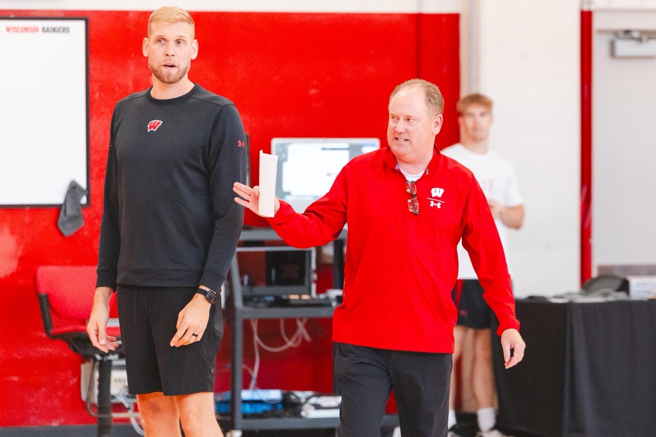wisconsin basketball coach greg gard in a red shirt and black pants holds a sheet of paper while pointing to direct players