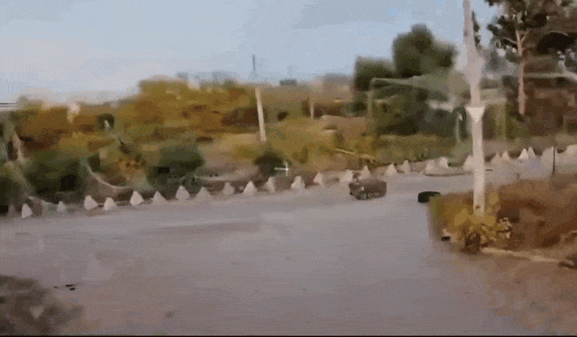 A small military-style vehicle drives into a flooded intersection and is quickly swept sideways by strong rushing water.