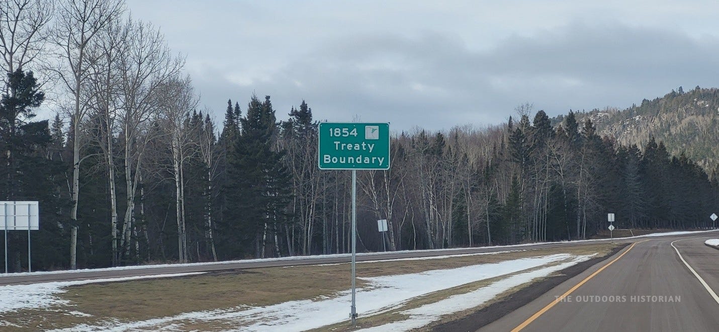 A brown roadside sign along a highway in Minnesota that reads "1854 Treaty Boundary." The background shows the edge of a boreal forest under a grey sky.