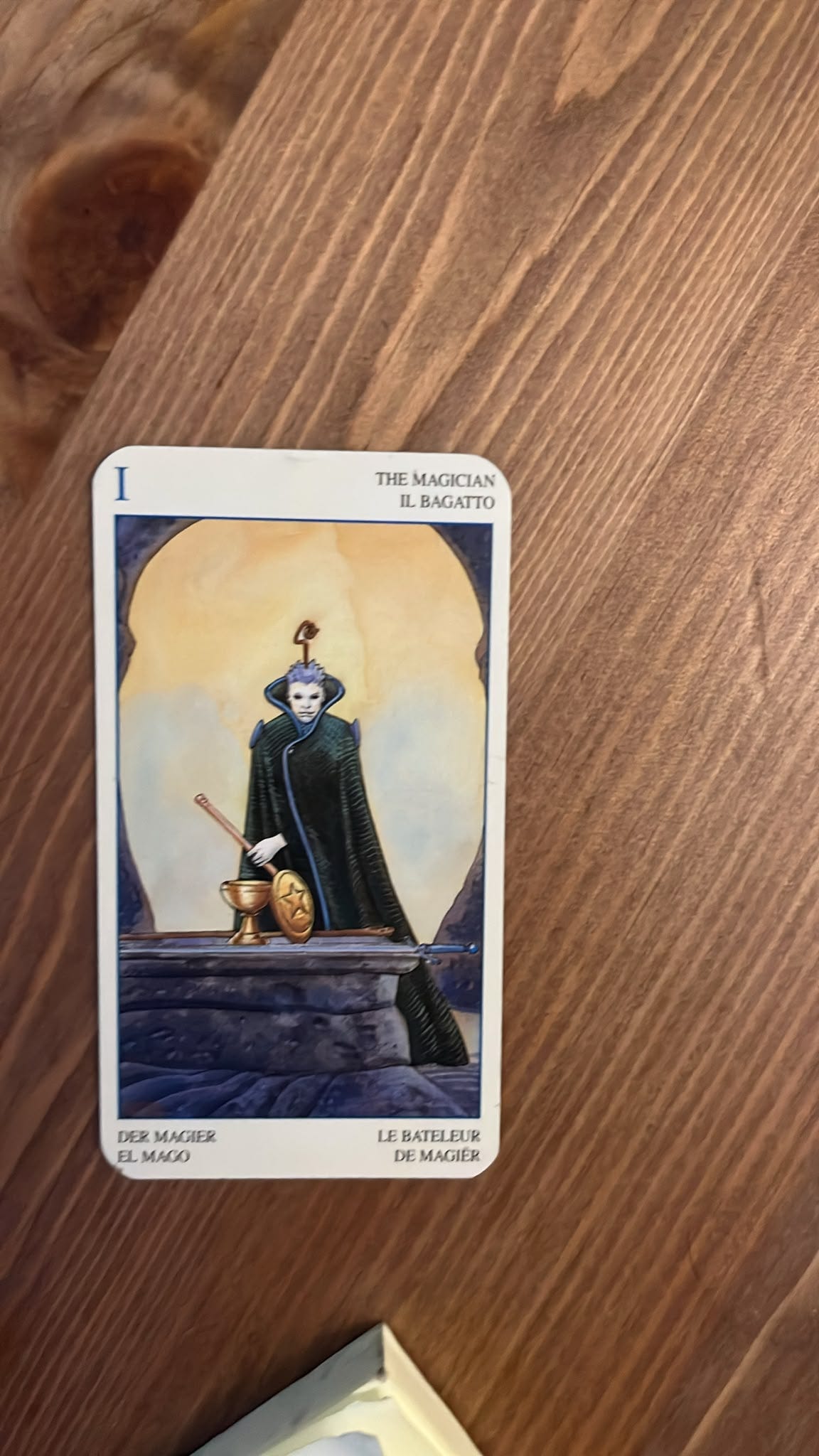 The Magician tarot card lies on a wooden table, depicting a pale, dark-robed figure standing behind a stone altar with a wand, cup, disc and sword. The Magician tarot card lies on a wooden table, depicting a pale, dark-robed figure standing behind a stone altar with a wand, cup, disc and sword.