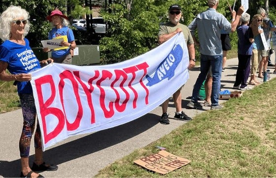 Photo: A large group of activists hold various signs and a large BOYCOTT AVELO banner outside the Cherry Capital Airport