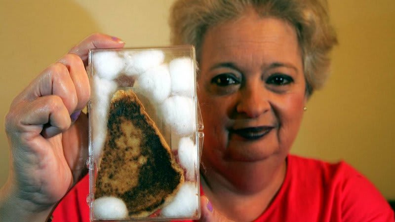 Lady with grey hair holding up a grilled cheese sandwich encased in glass with what seems to be a woman's face burnt into it - the "Holy Toast"