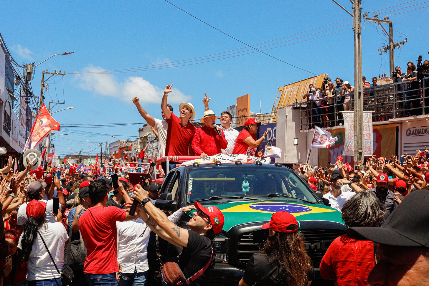 Multidão participa de uma manifestação política em uma rua movimentada, sob o sol. No centro da imagem, o presidente Luiz Inácio Lula da Silva, vestido de vermelho e usando chapéu, está em pé sobre um veículo, acompanhado de apoiadores. Ao redor, manifestantes carregam bandeiras vermelhas do PT, da CUT e de movimentos sociais, além de cartazes e bandeiras de candidatos. Uma grande bandeira do Brasil cobre parte do capô do veículo. Multidão participa de uma manifestação política em uma rua movimentada, sob o sol. No centro da imagem, o presidente Luiz Inácio Lula da Silva, vestido de vermelho e usando chapéu, está em pé sobre um veículo, acompanhado de apoiadores. Ao redor, manifestantes carregam bandeiras vermelhas do PT, da CUT e de movimentos sociais, além de cartazes e bandeiras de candidatos. Uma grande bandeira do Brasil cobre parte do capô do veículo.