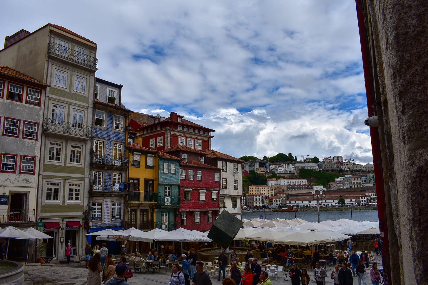 The colorful stacked facades of Ribeira Square along the Douro waterfront in Porto, Portugal, with outdoor restaurant seating and crowds along the cobblestone promenade.