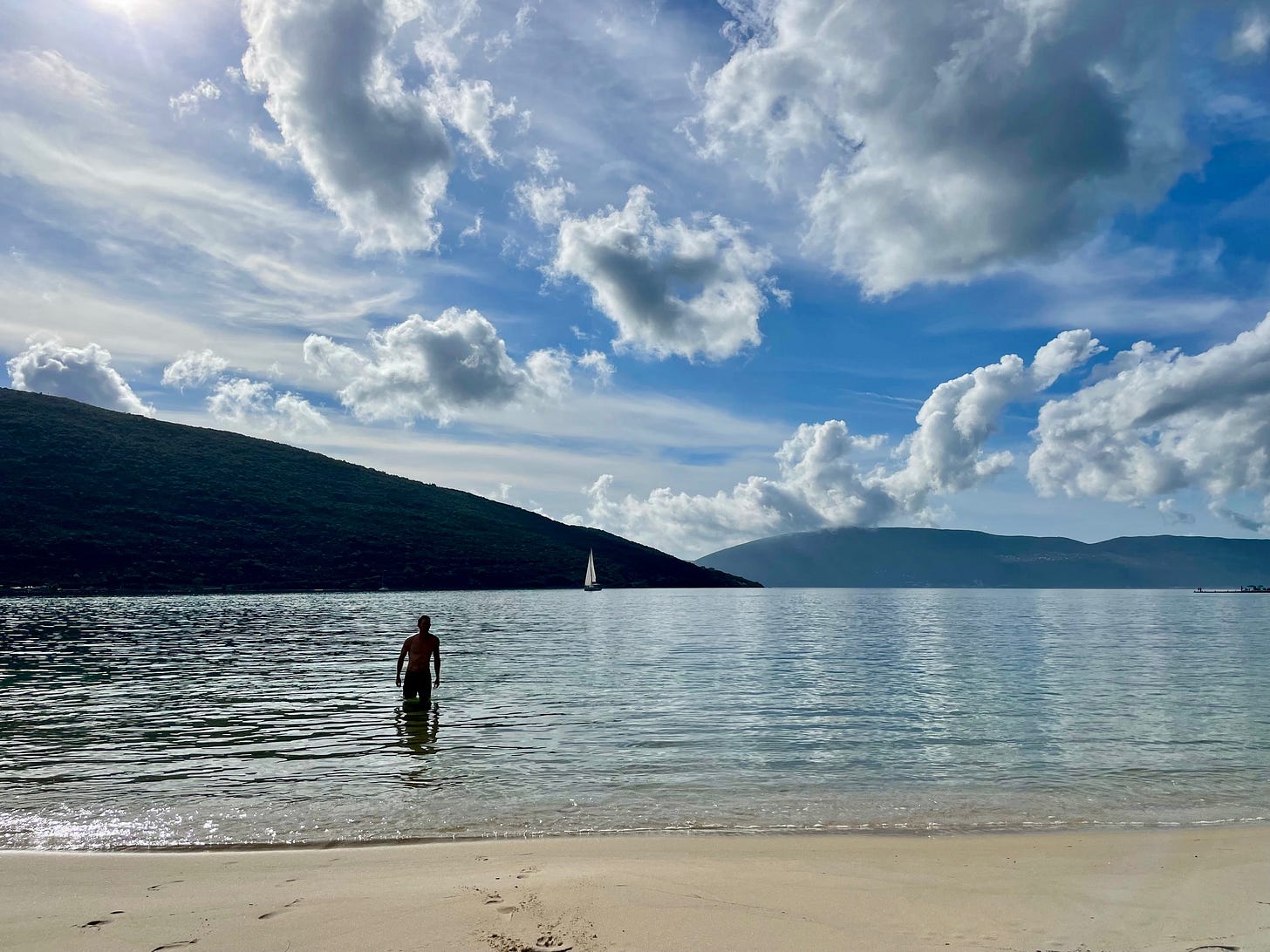 Person wading into the calm, clear water of a sandy Boka Bay beach in Montenegro, with a sailboat and dramatic mountain peaks in the background