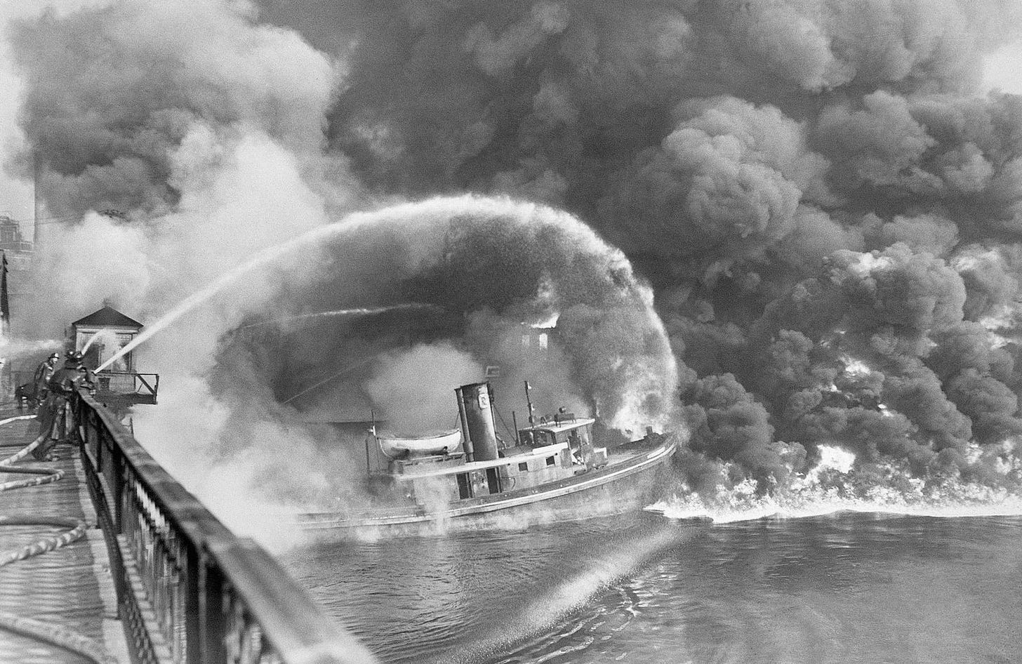 Firefighters spray water on burning boat amid heavy smoke.