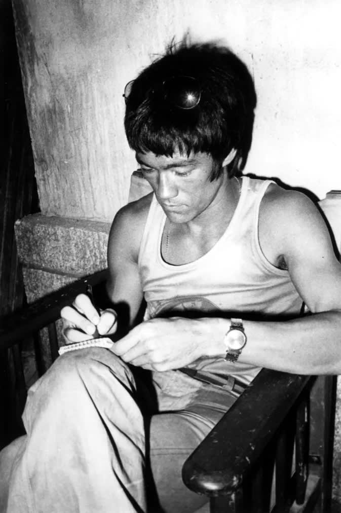 black and white photo of Bruce Lee sitting down and writing in a small notebook