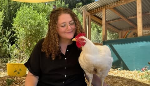 Curly-haired woman smiles and looks at a chicken on a farm