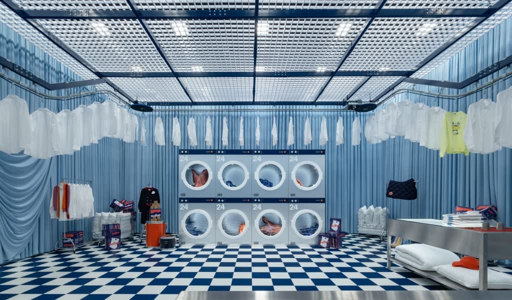 A whimsical laundry room in blue and white tones with checkered floors, curtained walls, and suspended shirts. Oversized dryers are filled with Hermès pieces and detergent boxes labeled “Galop.” Photograph by Sui Sicong. Courtesy of Hermès. A whimsical laundry room in blue and white tones with checkered floors, curtained walls, and suspended shirts. Oversized dryers are filled with Hermès pieces and detergent boxes labeled “Galop.” Photograph by Sui Sicong. Courtesy of Hermès.