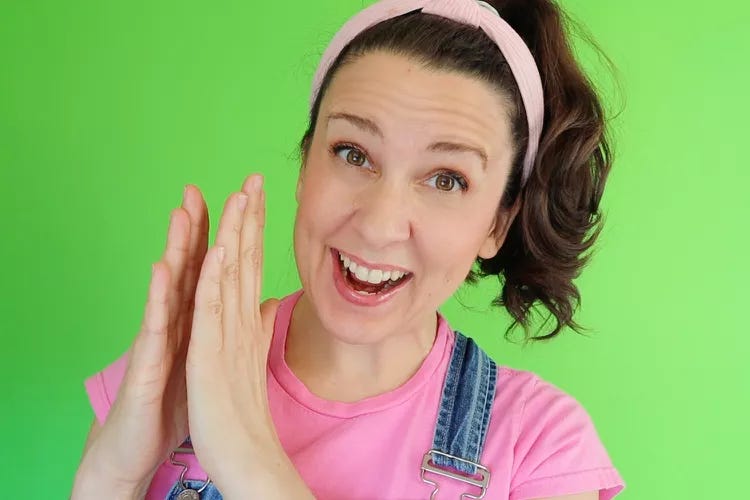 Picture of Ms. Rachel in a pink shirt, pink headband, and blue overalls. She's mid-clap, or perhaps PUTTING HER HANDS TOGETHER TO ALERT A WARLORD TO HER WHEREABOUTS.