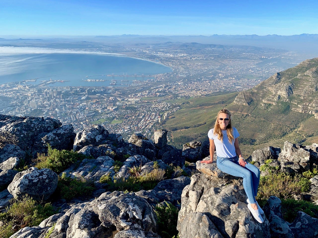 A woman sitting on a rocky outcrop high above Cape Town with the entire city, harbor, and coastline spread out far below her.