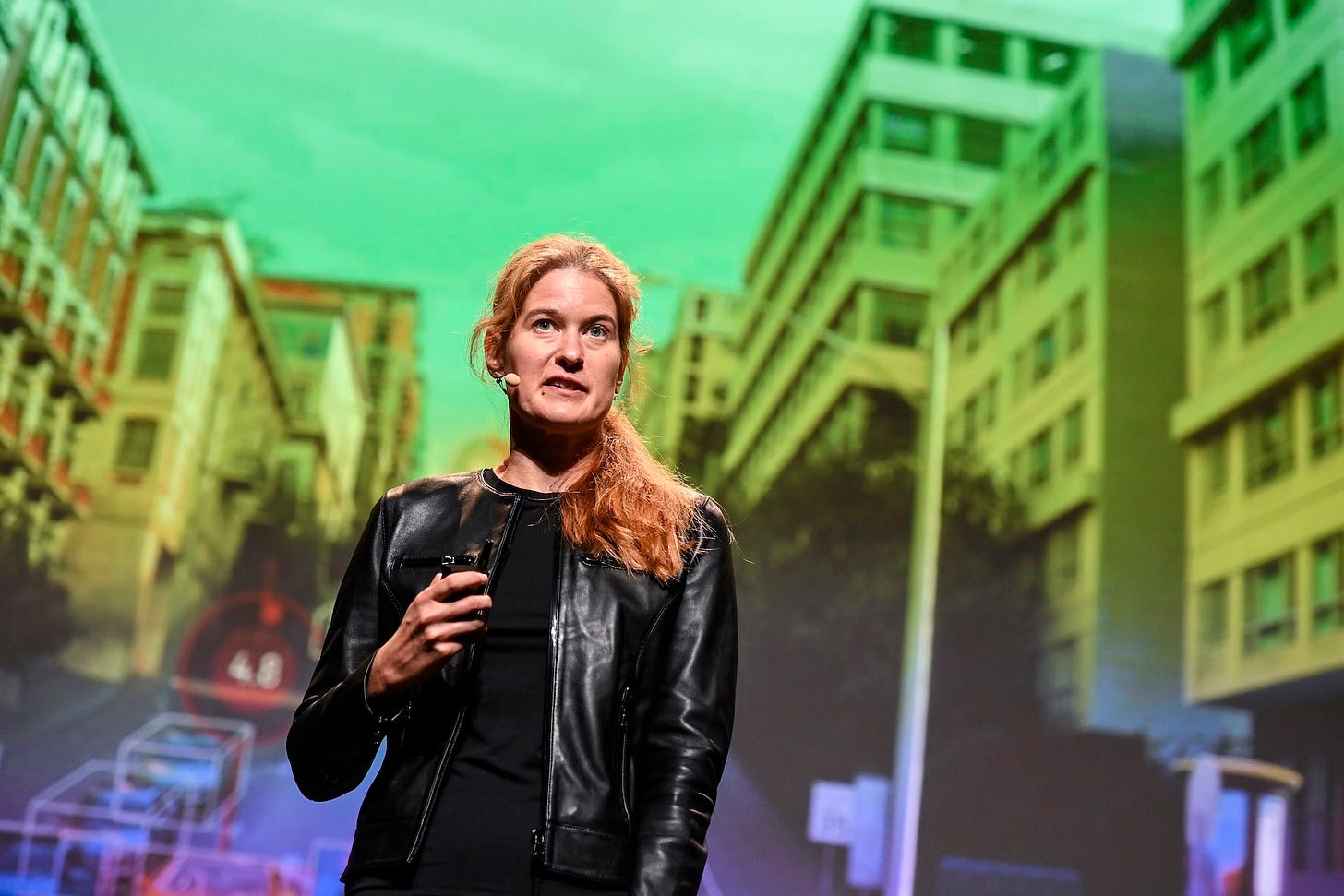 A woman wearing a ponytail and a black leather jacket stands on a stage. She looks slightly away from the camera. She wears a microphone and appears to be speaking to an audience.