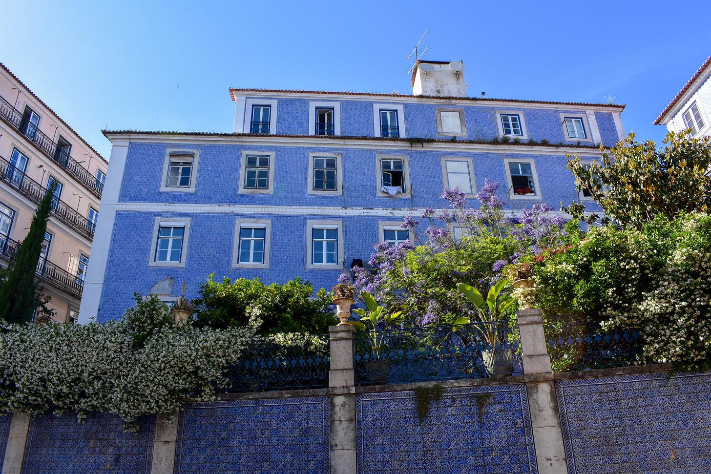 A traditional blue azulejo-tiled apartment building in Lisbon with white-trimmed windows and purple bougainvillea climbing a garden wall in front
