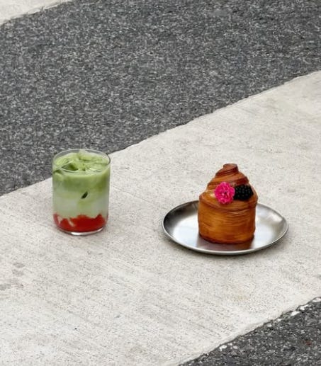A green matcha drink with a layer of red syrup beside a flaky stuffed pastry topped with blackberries and a pink edible flower, on a sidewalk curb near Etma bakery in Lisbon