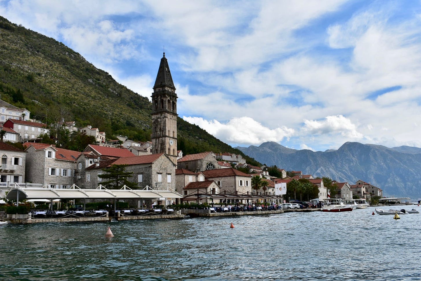 Perast's stone waterfront and 17th-century church bell tower seen from the water in Boka Bay, Montenegro, with terracotta-roofed buildings and mountains rising behind the village