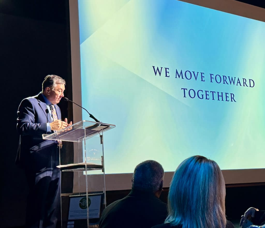 Mayor Ben Clapsaddle stands at a clear podium on stage, speaking into a microphone. A large projection screen behind him displays the text "WE MOVE FORWARD TOGETHER" against a blue and green gradient background. Mayor Ben Clapsaddle stands at a clear podium on stage, speaking into a microphone. A large projection screen behind him displays the text "WE MOVE FORWARD TOGETHER" against a blue and green gradient background.