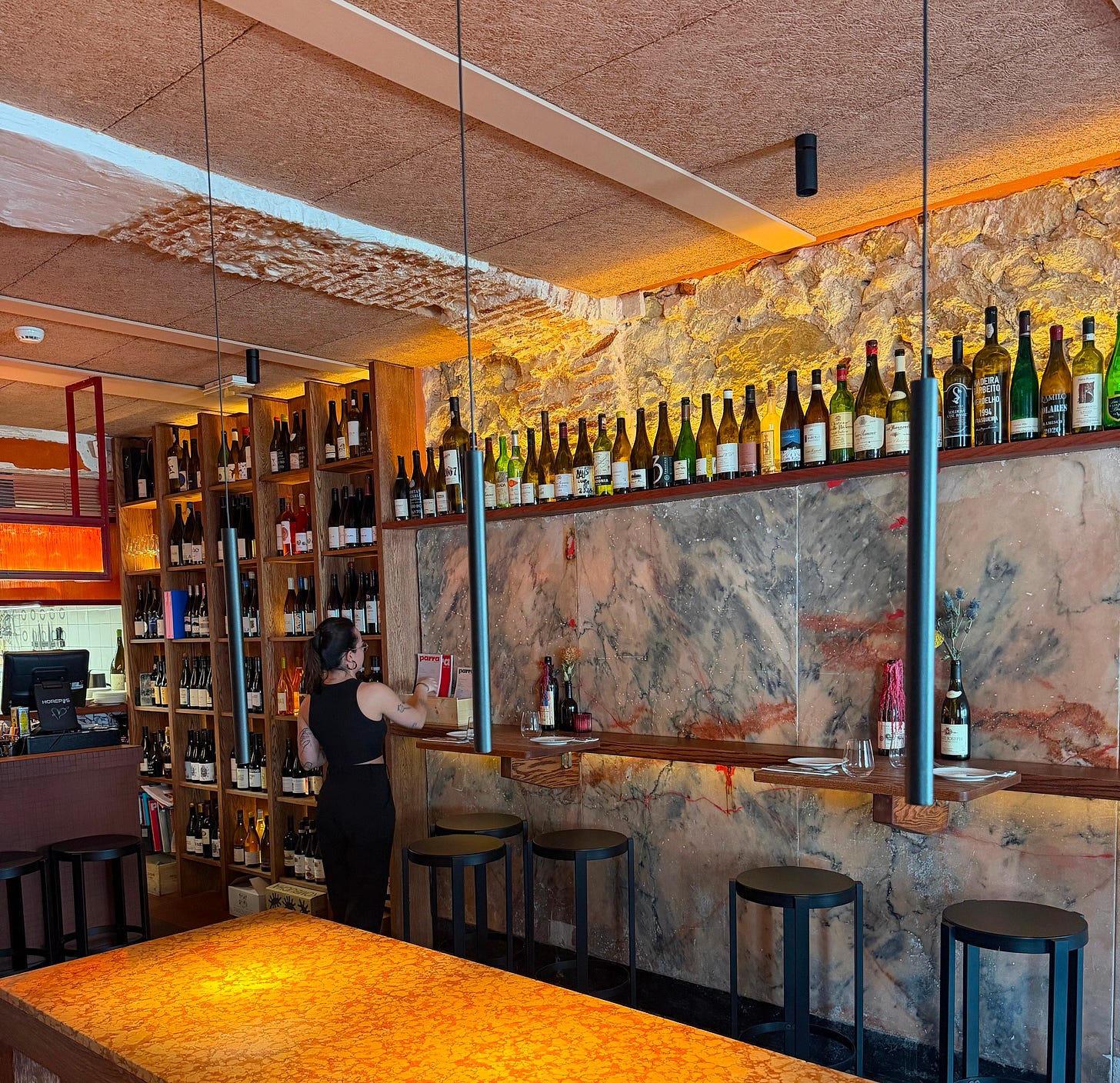 The interior of Parra Wine Bistro in Lisbon, with a marble accent wall, hanging pendant lights, and a server arranging wine bottles on open shelving