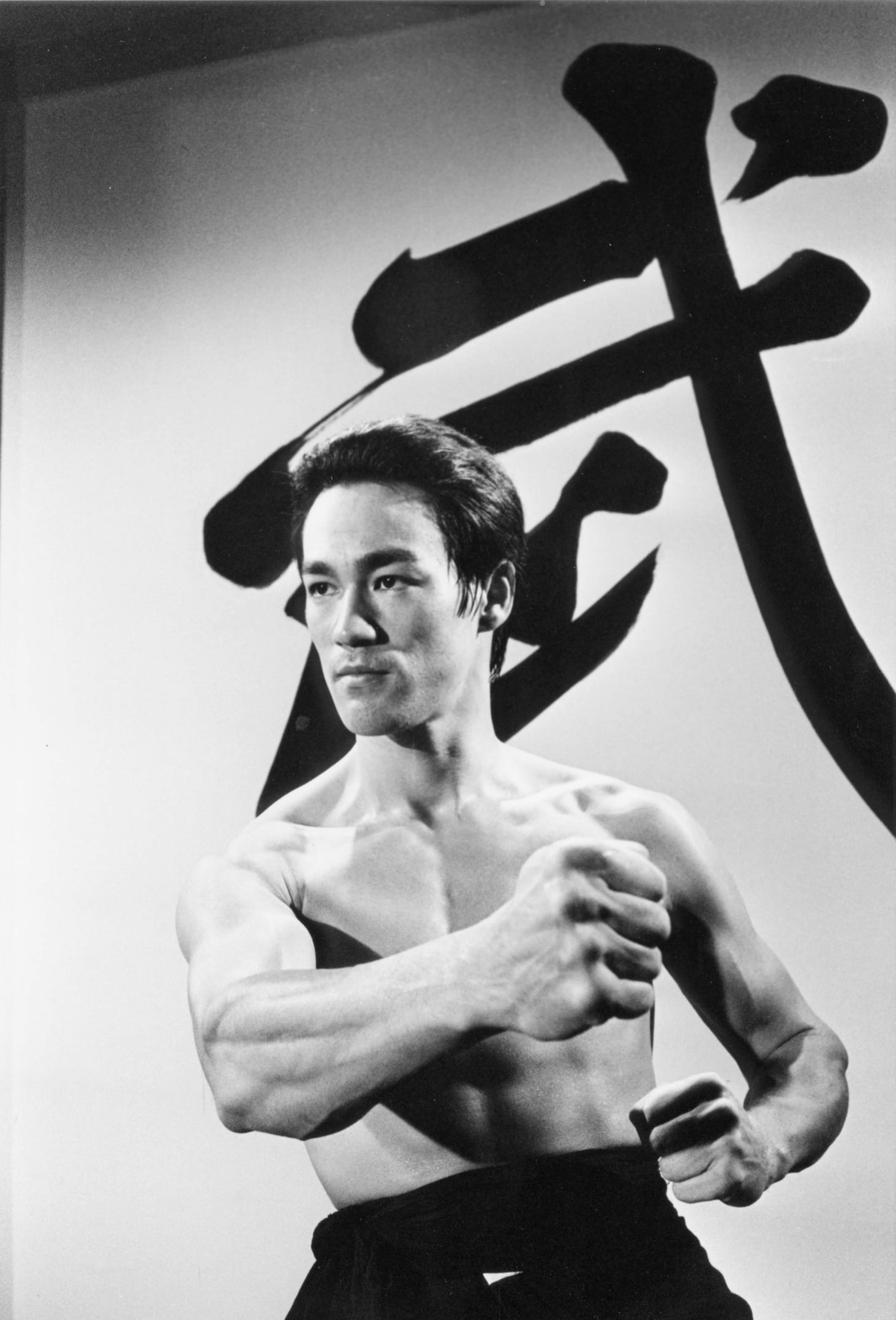 black and white portrait of Bruce Lee, shirtless, posing athletically with his fist extended, a large Chinese character looms on the backdrop
