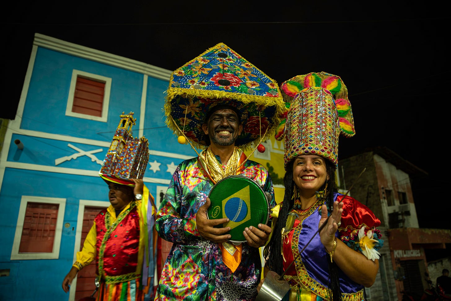 Três pessoas vestidas com trajes tradicionais coloridos e chapéus ornamentados participam de uma manifestação cultural popular à noite. Em primeiro plano, um homem sorridente segura um pandeiro com a bandeira do Brasil, ao lado de uma mulher que sorri e gesticula com as mãos. Ao fundo, outra pessoa também usa roupas festivas. A cena acontece em uma rua com casarões históricos, incluindo uma fachada azul. Três pessoas vestidas com trajes tradicionais coloridos e chapéus ornamentados participam de uma manifestação cultural popular à noite. Em primeiro plano, um homem sorridente segura um pandeiro com a bandeira do Brasil, ao lado de uma mulher que sorri e gesticula com as mãos. Ao fundo, outra pessoa também usa roupas festivas. A cena acontece em uma rua com casarões históricos, incluindo uma fachada azul.