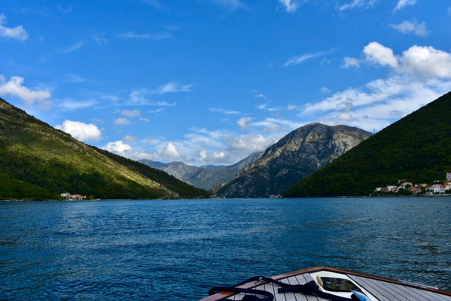 View from the bow of a private wooden boat crossing Boka Bay in Montenegro, surrounded by steep forested mountains under a wide blue sky