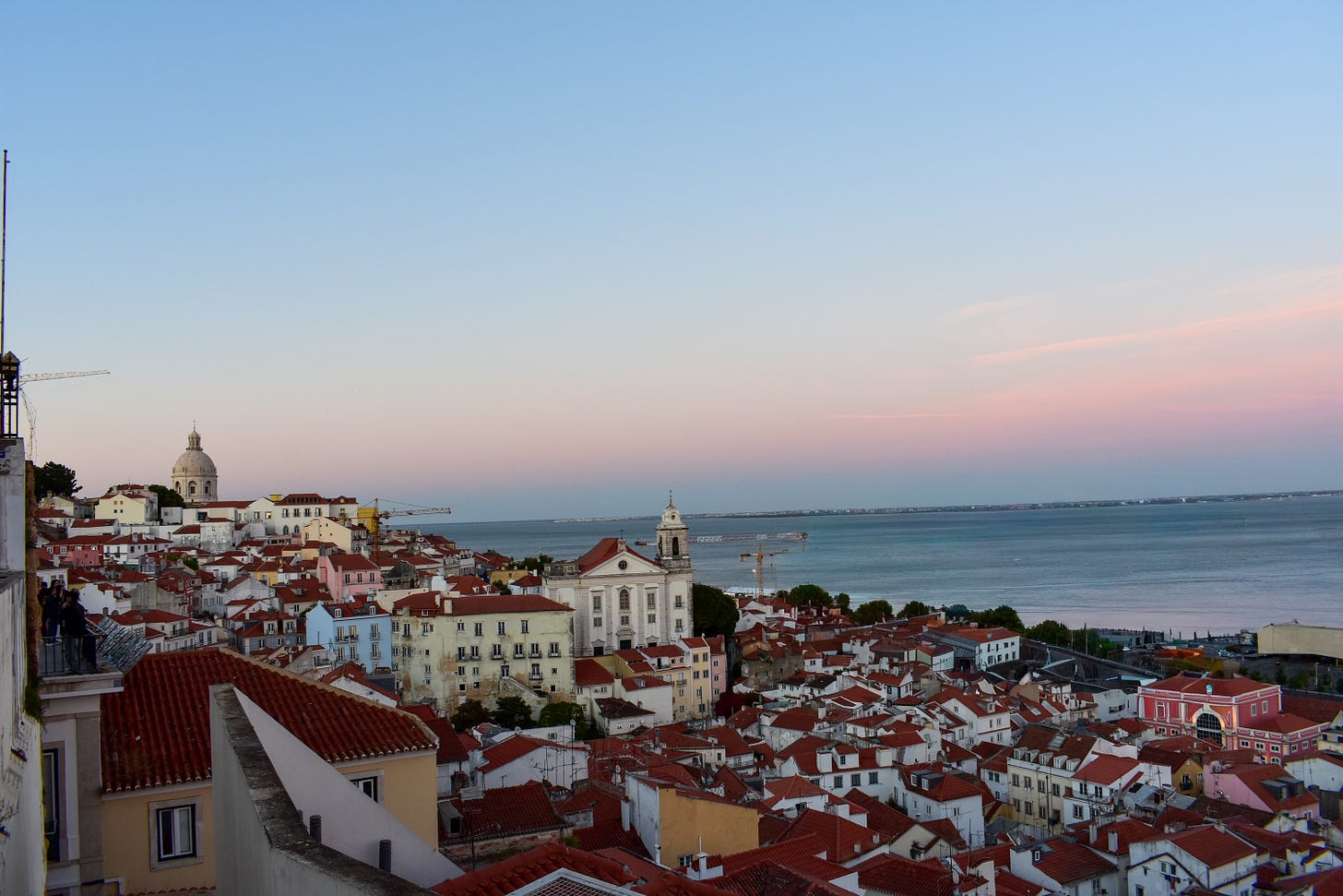 View over Alfama's red-tiled rooftops in Lisbon at sunset, with the dome of the National Pantheon and the Tagus River in the background