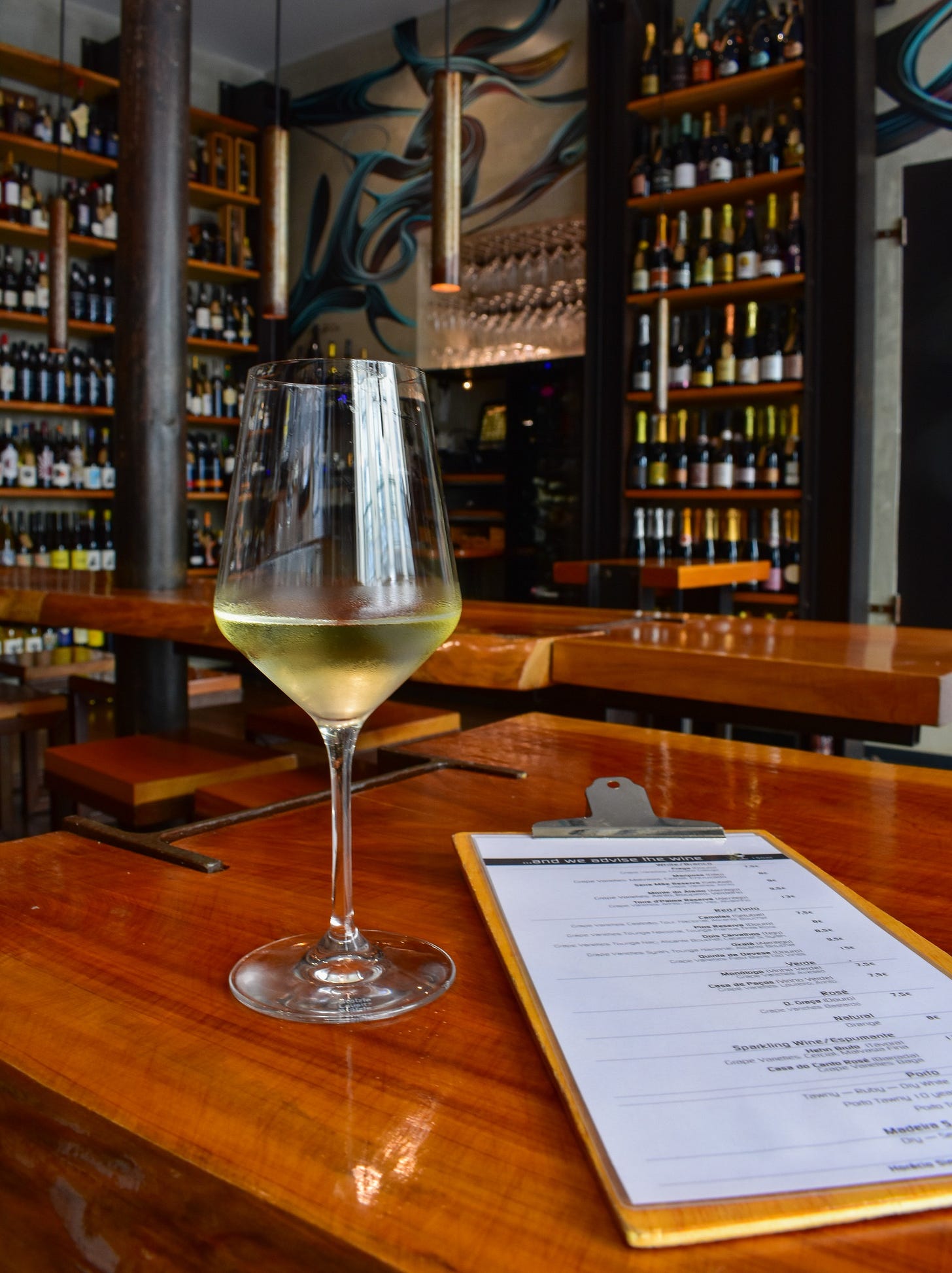 A glass of white wine and a handwritten wine list on a wooden bar at Nova Wine Bar in Lisbon, with floor-to-ceiling bottle shelves in the background