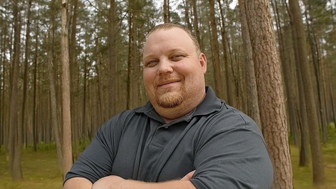 Brandon Huber, a Montana pastor and Realtor, stands outdoors with his arms crossed in a wooded area, smiling calmly after winning a long legal fight over religious freedom.