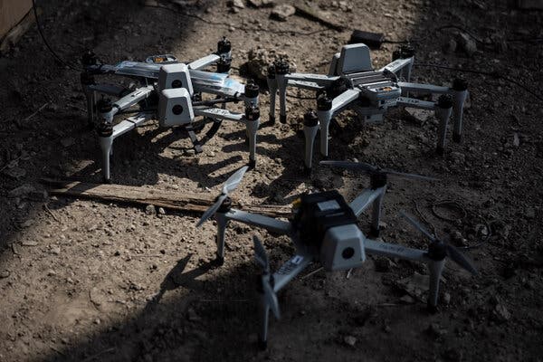 Three small military attack drones sit on a patch of dirt with a splash of sunlight highlighting the middle of the frame.