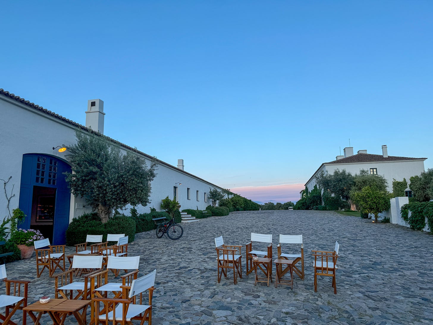 The main cobblestone courtyard at dusk, with white canvas chairs arranged for cocktail hour, olive trees, and a pale pink sunset sky visible between the whitewashed buildings