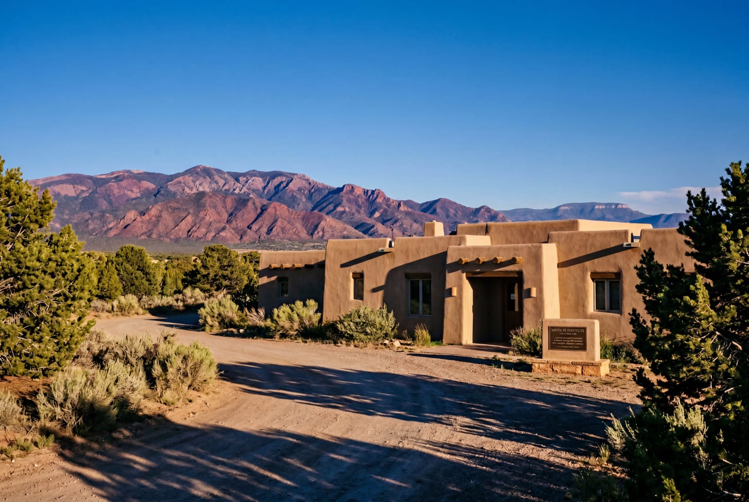 A late-afternoon exterior photograph of a low-slung adobe-style institutional research building in the Santa Fe, New Mexico high-desert landscape, framed by pi&ntilde;on pines and sage. The Sangre de Cristo mountains rise in the middle distance under a cobalt sky, and a distant mesa silhouette suggests the Los Alamos ridge on the far horizon. A small bronze plaque near the entrance reads 