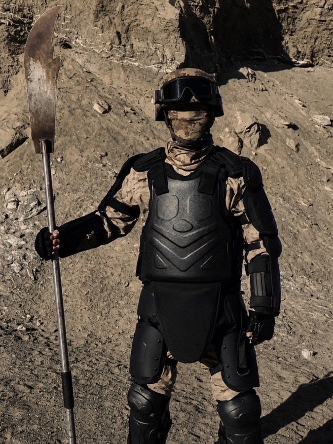 Armored soldier in camouflage holding a large blade on rocky terrain.