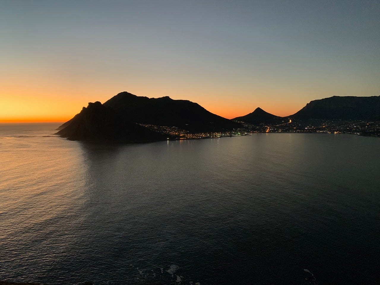 Mountains silhouetted against an orange and pink sunset, with calm water reflecting the sky and a small town visible along the coastline.