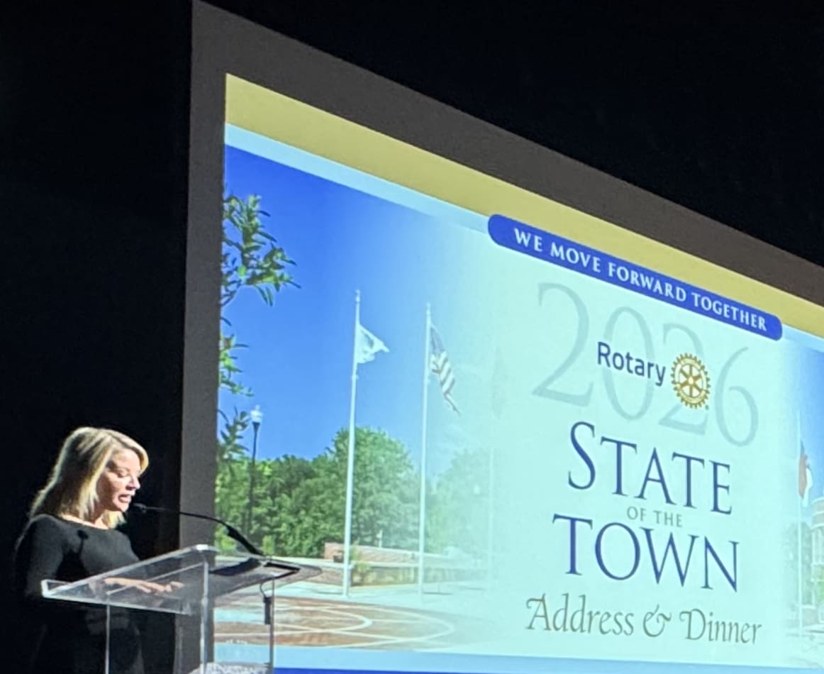 Angela Taylor stands at a clear podium on a dark stage, speaking into a microphone. Behind her, a large projection screen displays the Rotary logo and the event title, "2026 State of the Town Address & Dinner," beneath a banner reading "We Move Forward Together." Angela Taylor stands at a clear podium on a dark stage, speaking into a microphone. Behind her, a large projection screen displays the Rotary logo and the event title, "2026 State of the Town Address & Dinner," beneath a banner reading "We Move Forward Together."