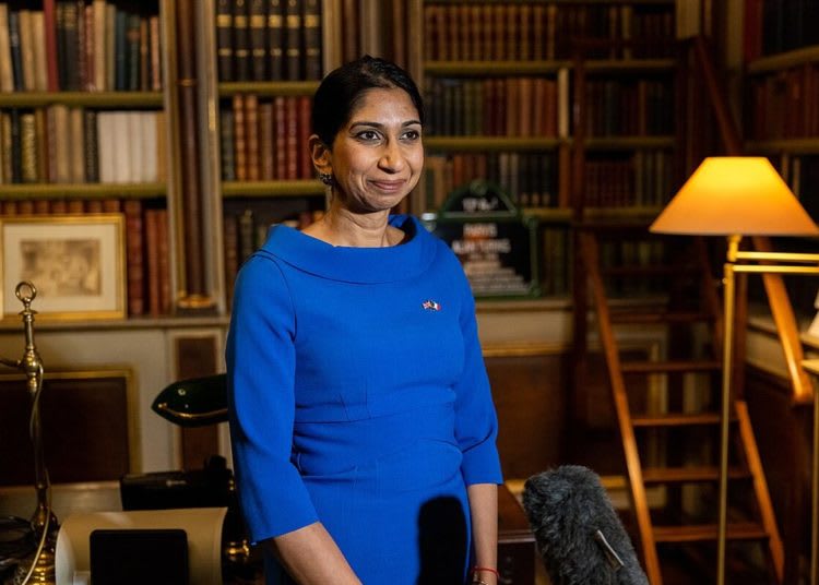 A portrait photo of Suella Braverman, then Home Secretary for the Conservative Party, in a library with wooden shelves and a switched on standard lamp to the right.