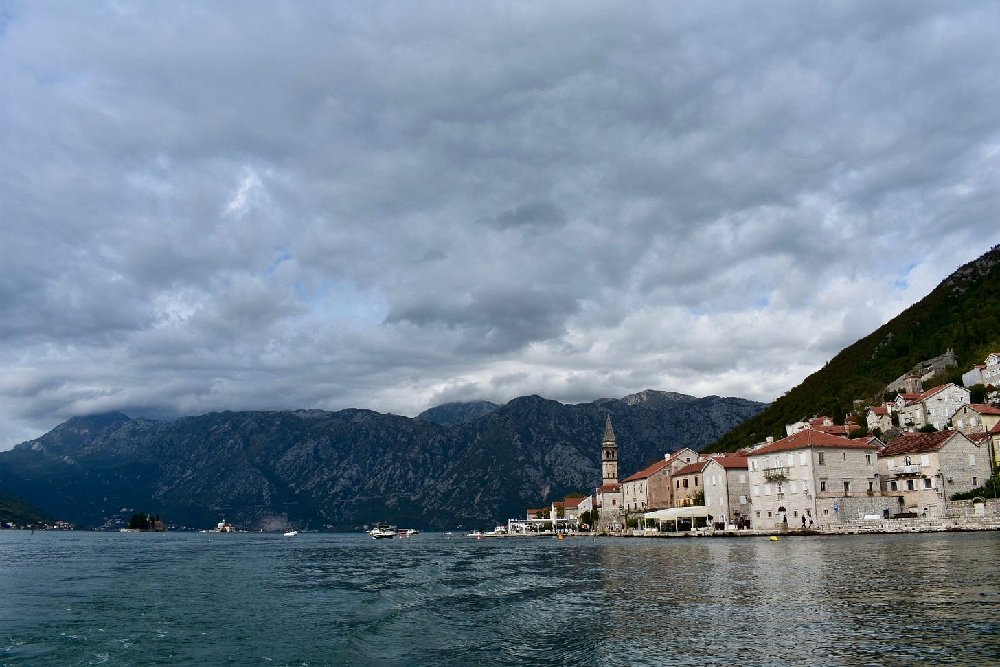 View from the water of Perast's Venetian waterfront, Montenegro, with stone palaces lining the bay, a church bell tower, and Our Lady of the Rocks island visible in the distance