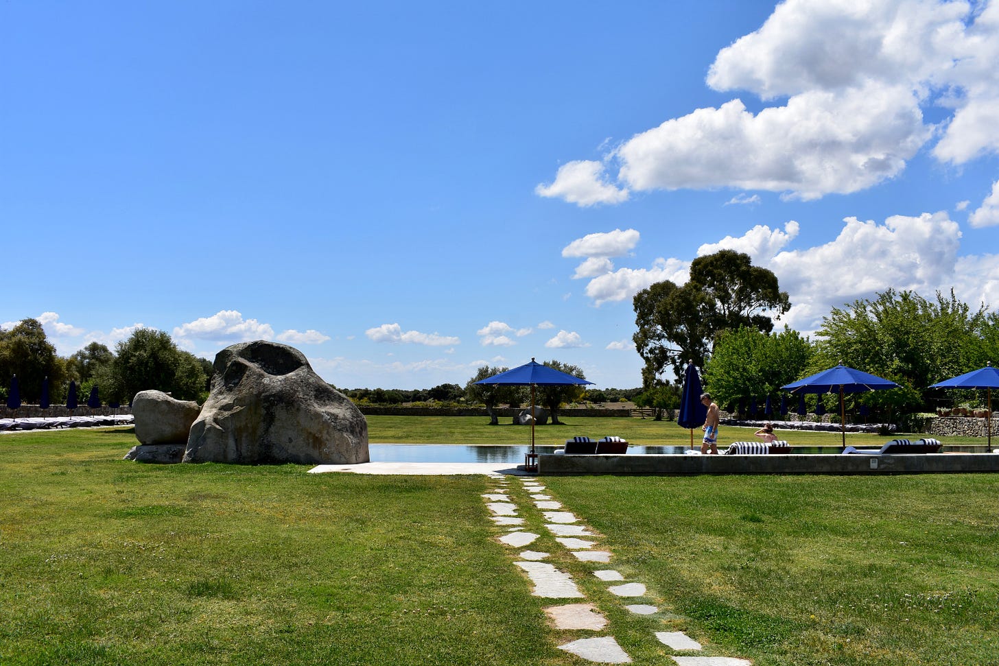 The outdoor pool at São Lourenço do Barrocal, Alentejo, with large natural granite boulders, navy umbrellas, and a stone stepping path across a wide green lawn