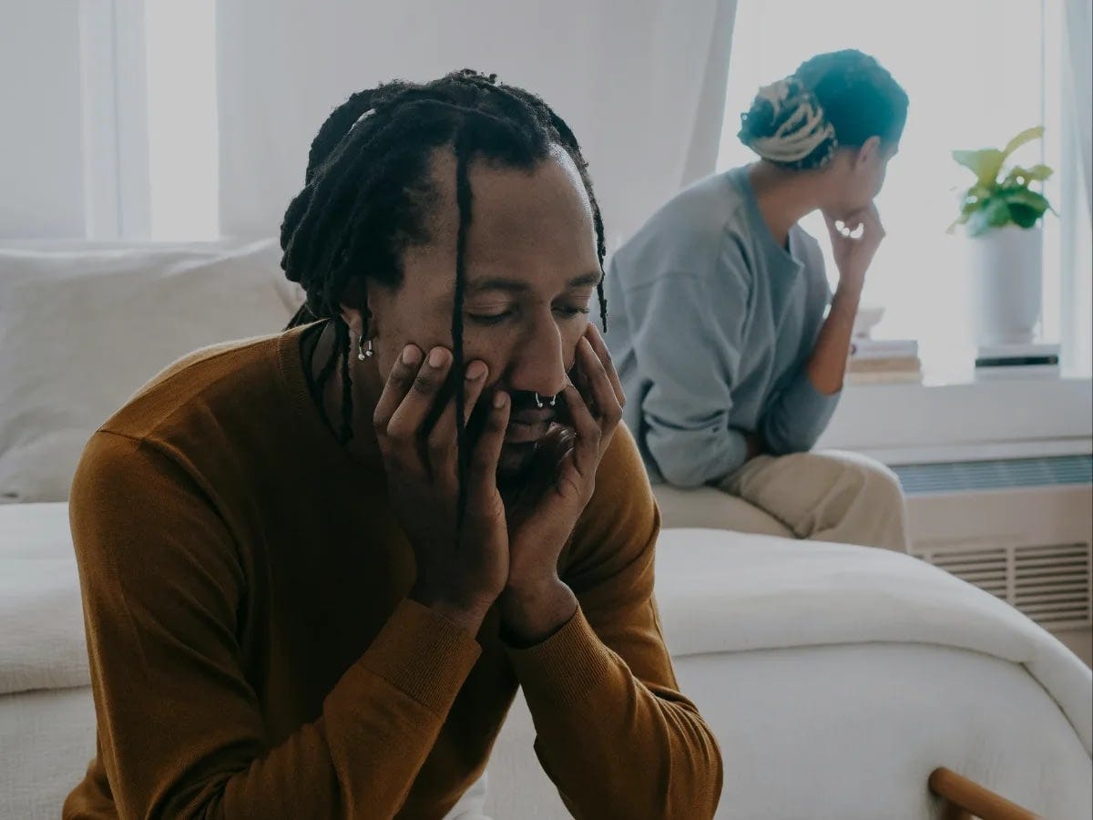 Man with dreadlocks in a brown sweater sits close to the camera, hands on his face, looking distressed.