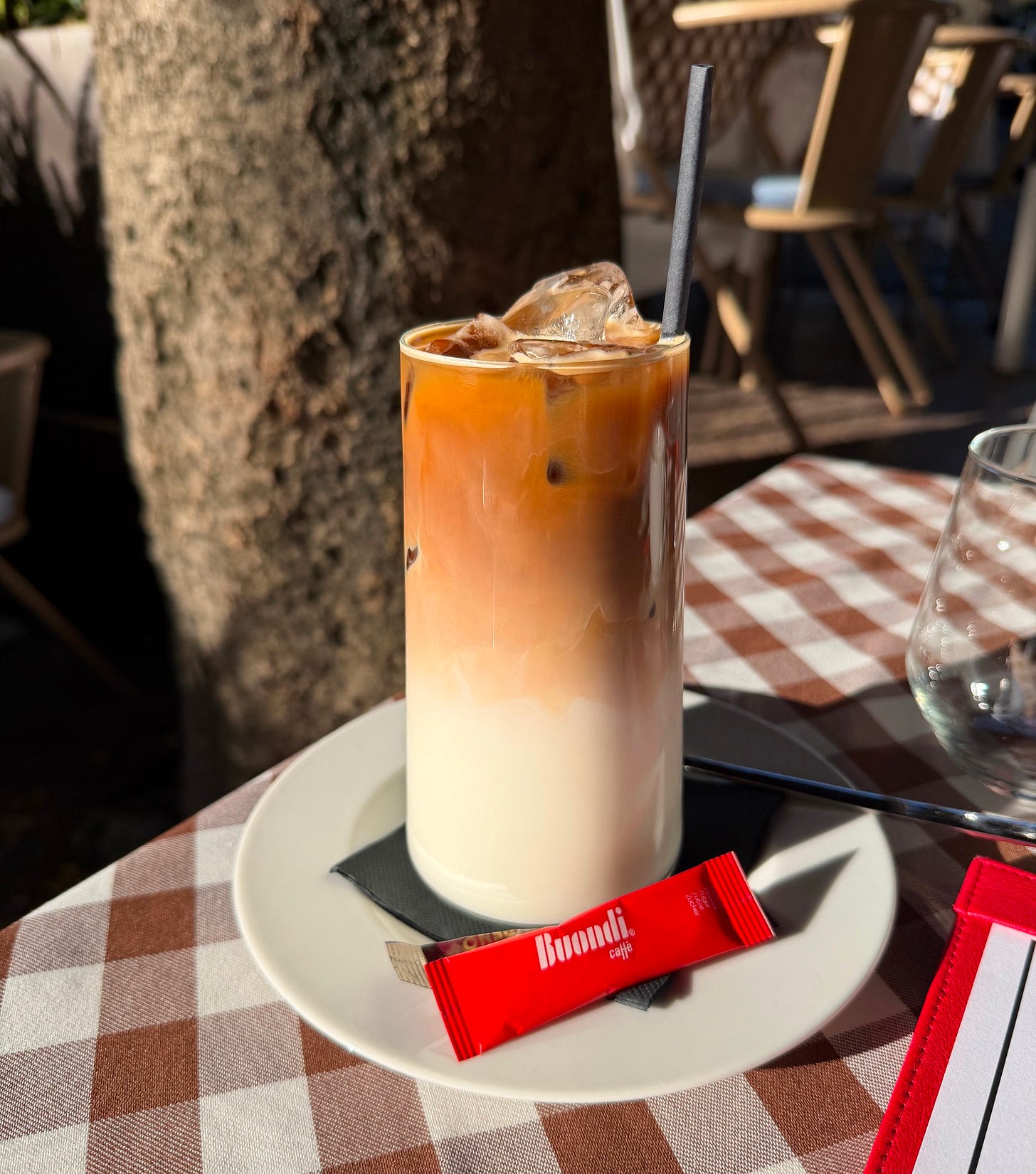 An iced coffee with a layered caramel color on a white plate, served on a checkered tablecloth at an outdoor cafe table in Lisbon